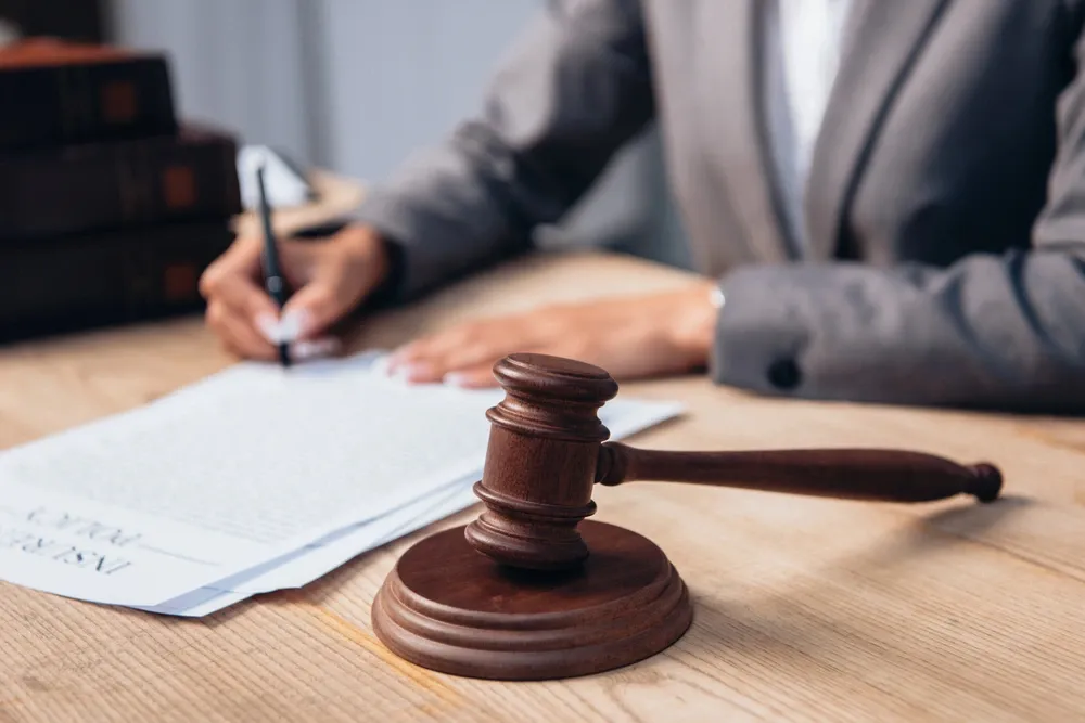 Close-up of a judge’s gavel on a desk with a person in a suit writing on legal documents in the background.